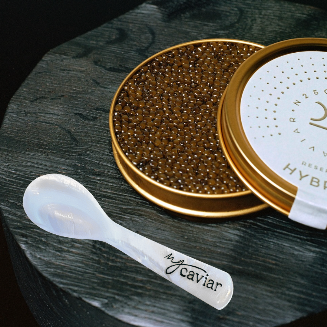 Caviar in a gold container with a white lid on a dark surface, accompanied by a caviar spoon.